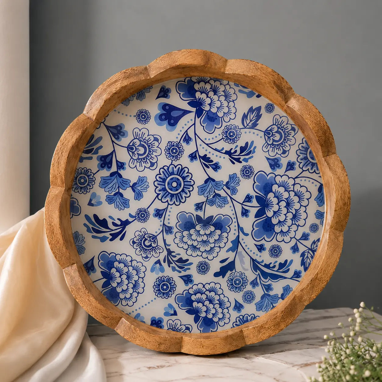 A round wooden serving tray with a scalloped edge, featuring a blue and white floral pattern on the inside, styled on a marble surface with a draped cream fabric and a gray background.