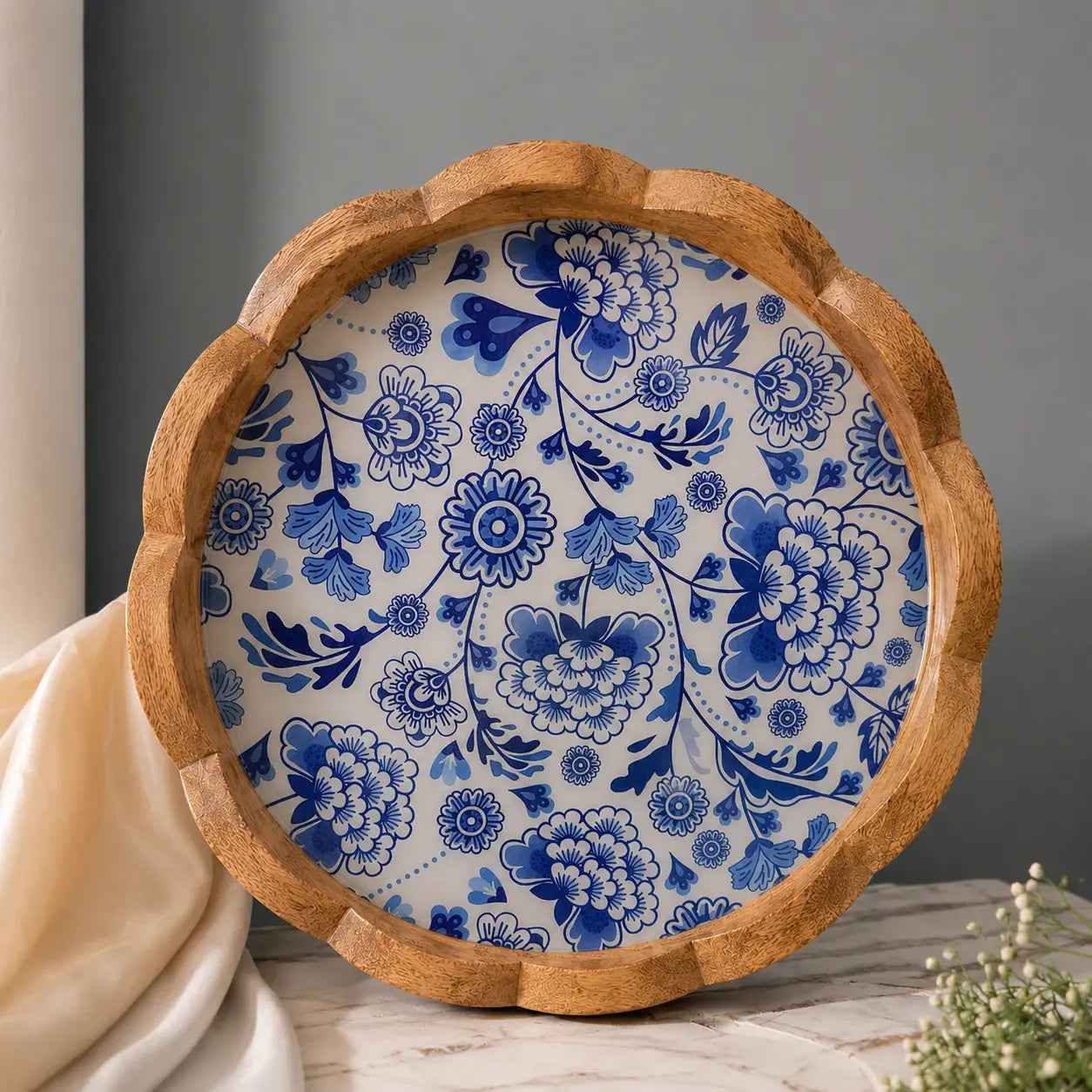 A round wooden serving tray with a scalloped edge, featuring a blue and white floral pattern on the inside, styled on a marble surface with a draped cream fabric and a gray background.