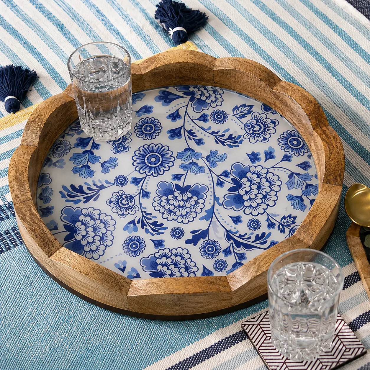 A round wooden serving tray with a scalloped edge, featuring a blue and white floral pattern on the underside, styled on a blue and white striped blanket with two clear glasses on it.