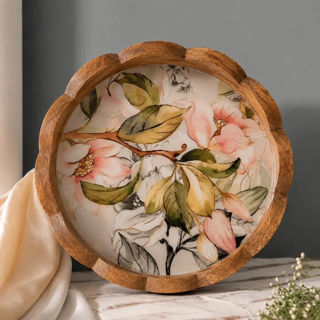 Round wooden decorative tray with a scalloped edge, featuring a hand-painted floral design of pink blossoms and green leaves on a cream background, styled on a marble surface with draped fabric.