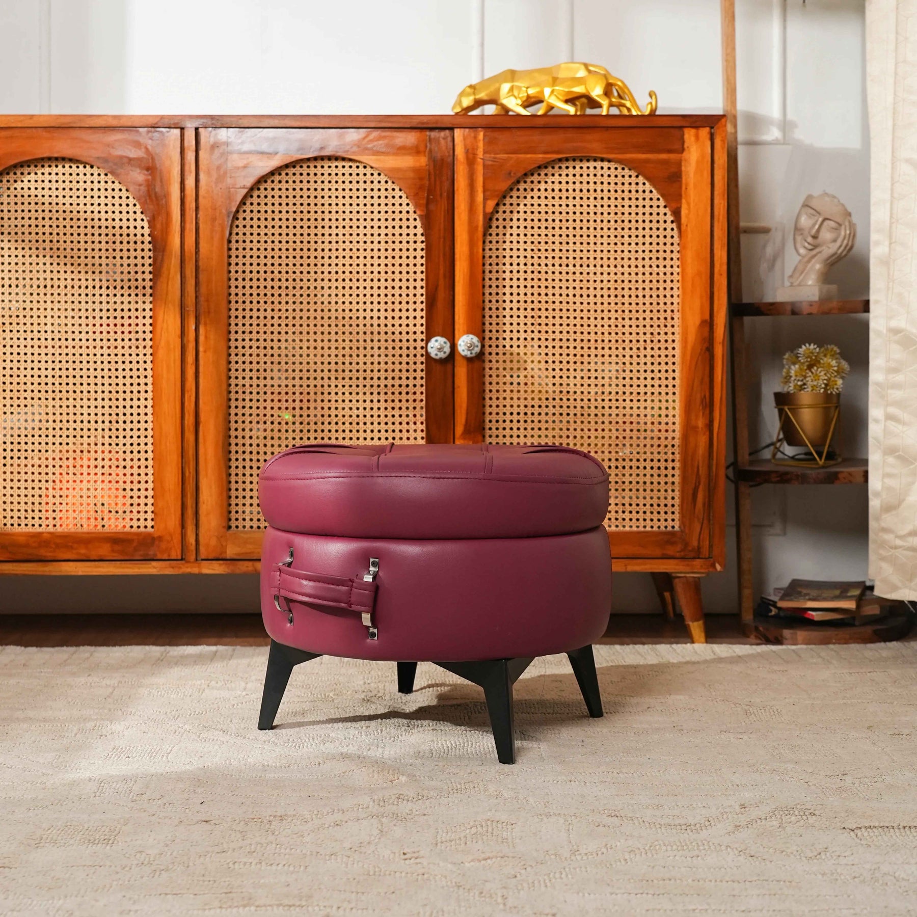 Maroon ottoman with suitcase design in front of a wooden cabinet.