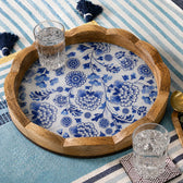 A round wooden serving tray with a scalloped edge, featuring a blue and white floral pattern on the underside, styled on a blue and white striped blanket with two clear glasses on it.