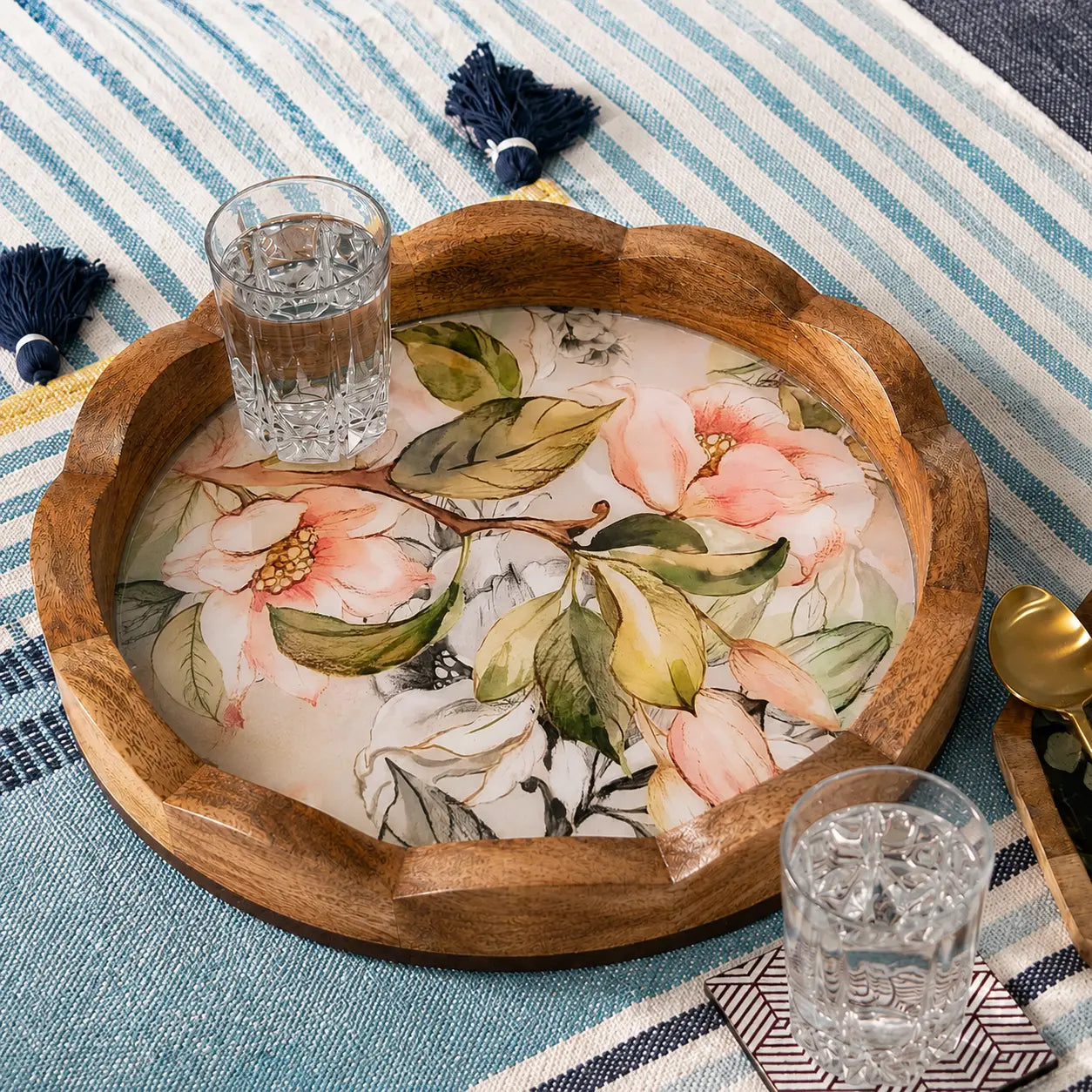 A scalloped wooden serving tray with a floral print, styled on a blue and white striped blanket with glasses, a spoon, and tassels.